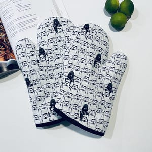 May include: White oven mitts with a black and white Star Wars Stormtrooper and Darth Vader pattern. The mitts have a dark blue trim. A cookbook and limes are in the background.