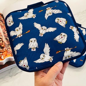 May include: Two square, navy blue pot holders with a white owl pattern. The owls are depicted with letters and brooms. The pot holders have a dark blue trim and a small loop for hanging.