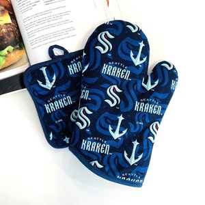 May include: A navy blue oven mitt and pot holder set. The set features a repeating pattern of the Seattle Kraken logo, anchors, and wave designs in white and light blue. The pot holder has a loop for hanging.