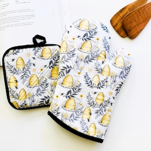 May include: A white oven mitt and pot holder set with a bee and honeycomb pattern. The design includes yellow honeycombs, bees, and floral elements on a white background. The set has black trim and a black loop on the pot holder.