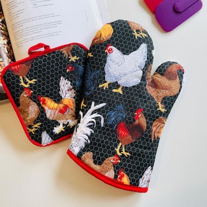 May include: A black oven mitt and a square pot holder featuring a repeating pattern of chickens and roosters in various colors, including white, brown, and red, set against a black chicken wire background with bright red fabric trim.