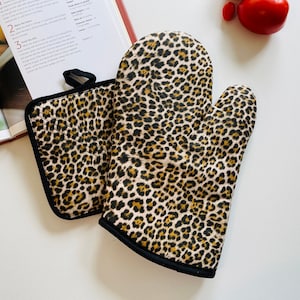 May include: A leopard print oven mitt and pot holder set. The mitt is a classic oven glove shape, and the pot holder is square. Both items feature a black trim and a loop for hanging. The pattern includes tan, brown, and black spots on a cream background.