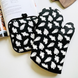 May include: Black oven mitt and pot holder set with a white ghost pattern. The kitchen textiles feature a repeating design of cartoon ghosts on a dark fabric. The set is next to an open cookbook.