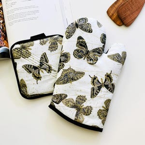May include: A white oven mitt and pot holder set with a black trim, featuring gold and black butterfly designs. The set is next to an open cookbook and wooden kitchen utensils.