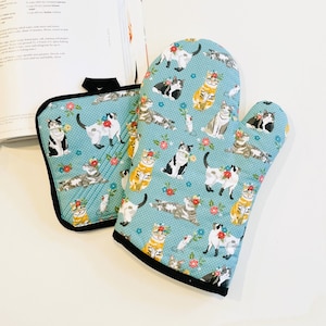 May include: A blue oven mitt and pot holder set with a cat and floral pattern. The cats are in various poses and colors, with red and pink flowers. The set has black trim and a quilted design.