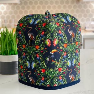 May include: A navy blue tea cozy with a vibrant tropical bird and floral pattern. The design features toucans, herons, and colorful flowers. The cozy has a rounded top and a dark blue trim.