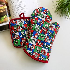 May include: A quilted oven mitt and pot holder set featuring a colorful pattern of Mario and Luigi characters in a grid design. The set has a red trim and a loop for hanging. The background includes a cookbook.