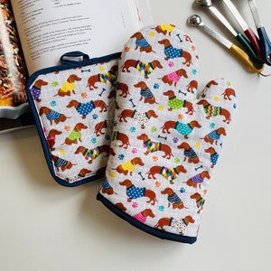 May include: A gray oven mitt and pot holder set with a navy blue trim. The set features a repeating pattern of dachshunds wearing colorful sweaters, paw prints, and bones. A cookbook and measuring spoons are in the background.