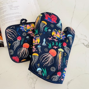 May include: A navy blue oven mitt and pot holder set. The set features a colorful jellyfish pattern with orange, yellow, pink, and blue accents. The design includes various sizes of jellyfish and aquatic plants.