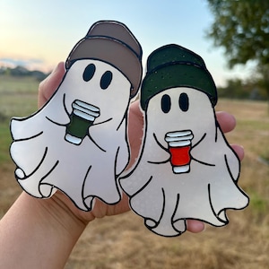 May include: Two ghost-shaped stickers, each holding a coffee cup. One ghost wears a brown hat and holds a green cup, the other wears a green hat and holds a red cup. The stickers have black outlines and are held in a hand.