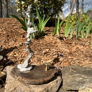 May include: A silver metal sculpture of a tree branch on a wooden slice with a gold metal mushroom on top. The sculpture is sitting on a rock in a garden setting.