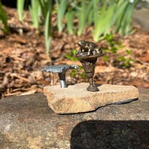 May include: A small bronze frog figurine perched atop a mushroom-shaped stand, both mounted on a grey rock. A second, smaller mushroom-shaped stand is also mounted on the rock.