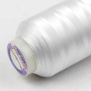 May include: Close-up of a white spool of WonderFil Deco Bob thread. The spool has a white plastic base with a purple and white label that reads "WonderFil DECO BOB DB-104". The thread is tightly wound and appears smooth and shiny.