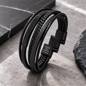 Peut inclure: Un bracelet noir multi-brins composé de cordons en cuir lisse, texturé et tressé. Il présente une barrette en métal noir mat et un fermoir magnétique gravé avec le texte Pursue Your Dreams KC PREMIUM sur la partie métallique.