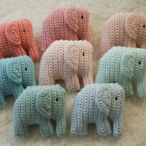 Könnte beinhalten: Sieben gehäkelte Elefantenfiguren in Pastellfarben: Koralle, Rosa, Hellrosa und zwei Blautöne. Jeder Elefant hat kleine schwarze Augen und wird auf einer weißen, strukturierten Oberfläche präsentiert.