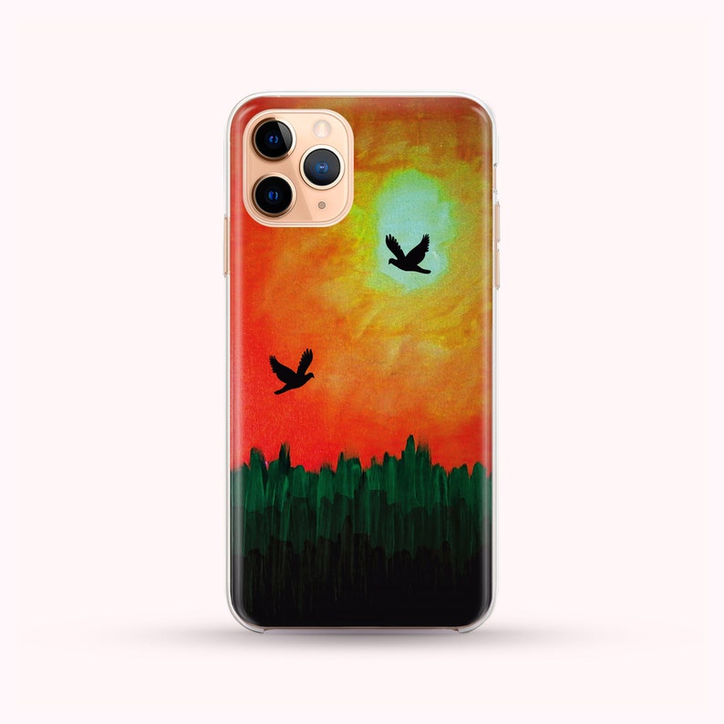 May include: A clear phone case with a watercolor sunset design. The sunset is painted in shades of orange, yellow, and red. Two black silhouettes of birds are flying in the sky. The bottom of the case is painted with a dark green forest.