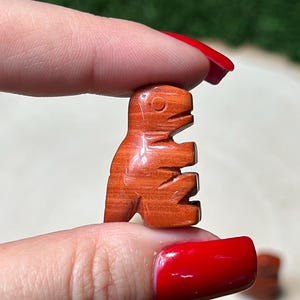 May include: A set of seven small, carved wooden dinosaur figurines. The dinosaurs are a reddish-brown color and are arranged in a circle on a wooden surface.