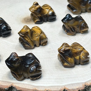 May include: A collection of small, carved frog figurines in shades of brown and black, displayed on a round wooden slice with a bark edge. The frogs are arranged on a light-colored surface, with green grass in the background.