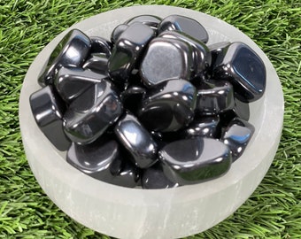 Puede incluir: Un cuenco lleno de piedras de hematites negras pulidas. Las piedras son lisas y brillantes, y están dispuestas en un patrón aleatorio. El cuenco es blanco y tiene una superficie texturizada.