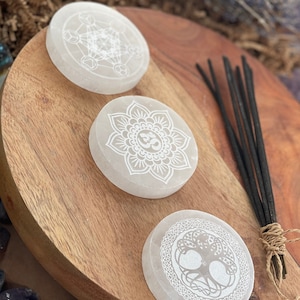 May include: Three white selenite coasters with different etched designs: a geometric pattern, a mandala with the Om symbol, and a tree of life. The coasters are on a wooden surface with a bundle of incense sticks.