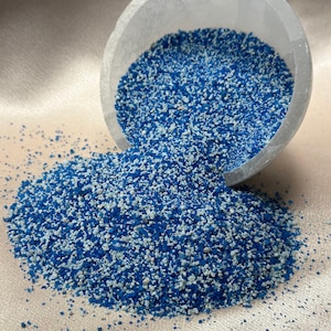 May include: A close-up of a white bowl filled with blue and white glitter. The bowl is tipped over, spilling the glitter onto a white surface.