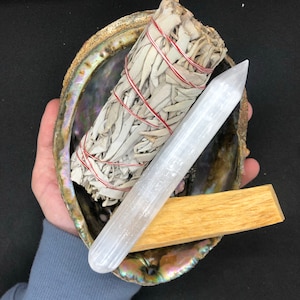 May include: A white sage smudge stick, a clear quartz crystal wand, and a palo santo stick are arranged in a large abalone shell.
