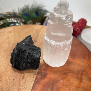 May include: A white selenite tower and a black obsidian crystal on a wooden surface. The selenite tower is tall and slender, with a pointed top. The obsidian crystal is rough and irregular in shape.