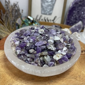 May include: A white selenite bowl filled with amethyst chips. The chips are various shades of purple and some have a white or clear appearance.