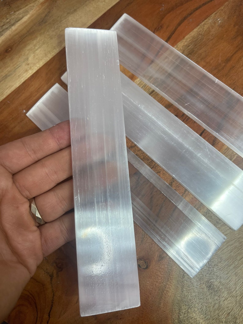 May include: Four selenite wands, also known as selenite sticks, are arranged on a wooden surface. The wands are smooth, white, and translucent. They are approximately 4 inches long and 1 inch wide.
