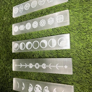 May include: Five white selenite charging wands with different etched designs. The top wand has seven chakra symbols, the second wand has seven eye symbols, the third wand has eight moon phases, the fourth wand has a moon phase with a star and the fifth wand has a moon phase with a globe.