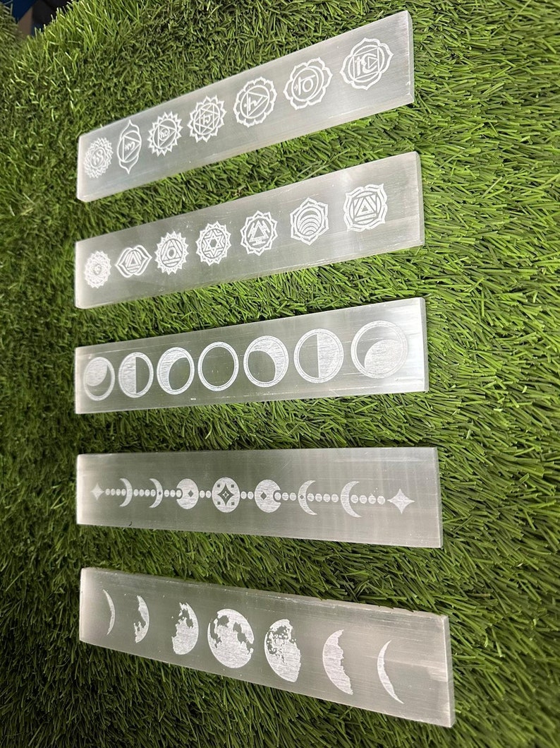 May include: Five white selenite charging wands with different etched designs. The top wand has seven chakra symbols, the second wand has seven eye symbols, the third wand has eight moon phases, the fourth wand has a moon phase with a star and the fifth wand has a moon phase with a globe.