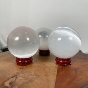 Op de afbeelding: Drie witte selenietkristallen bollen op rode houten bases. De bollen zijn glad en doorschijnend, met een lichte glans.