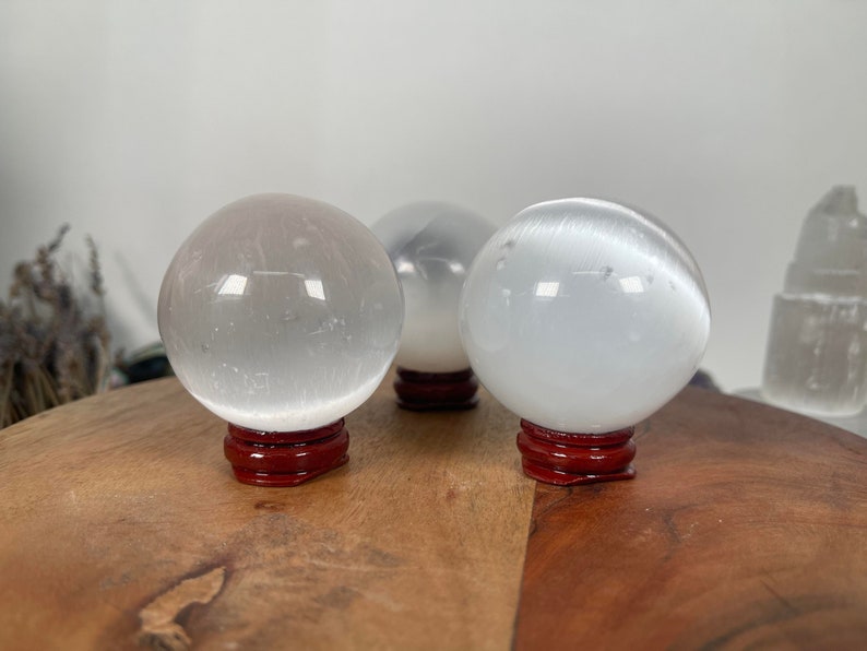 Op de afbeelding: Drie witte selenietkristallen bollen op rode houten bases. De bollen zijn glad en doorschijnend, met een lichte glans.