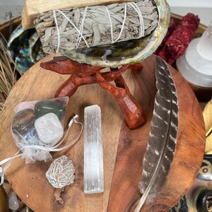 May include: A white sage smudge stick wrapped in twine and placed in a large abalone shell on a wooden stand. The shell is resting on a wooden tray with a feather, a selenite wand, a small bag of crystals, and a rose quartz crystal.