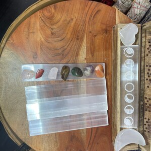 May include: A wooden tray with a selenite charging plate, a set of seven chakra stones, and a selenite heart and crescent moon. The tray is decorated with a moon phase design.