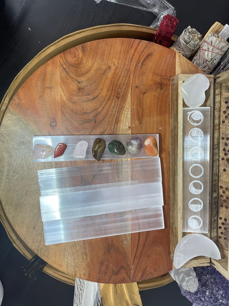 May include: A wooden tray with a selenite charging plate, a set of seven chakra stones, and a selenite heart and crescent moon. The tray is decorated with a moon phase design.
