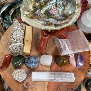 May include: A wooden tray with a variety of crystals, including a selenite wand, a rose quartz, a tiger's eye, and a clear quartz. There is also a sage bundle, a abalone shell, and a small white pouch.