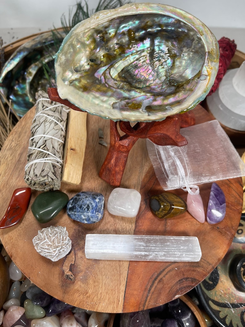 May include: A wooden tray with a variety of crystals, including a selenite wand, a rose quartz, a tiger's eye, and a clear quartz. There is also a sage bundle, a abalone shell, and a small white pouch.