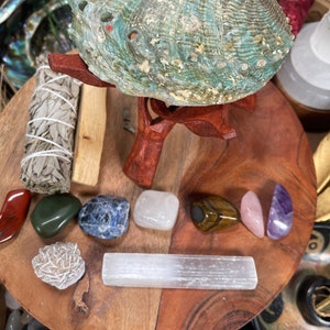 May include: A green abalone shell on a wooden stand surrounded by various crystals and stones, including selenite, amethyst, rose quartz, tiger's eye, and a sage smudge stick.