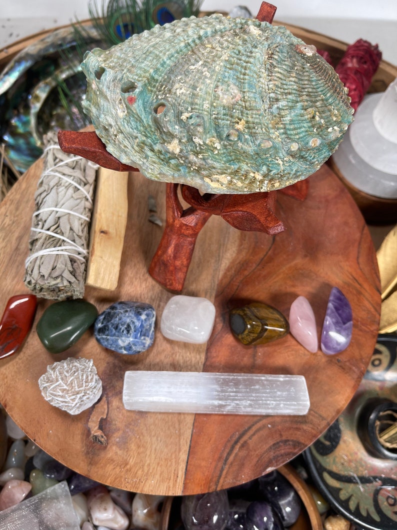 May include: A green abalone shell on a wooden stand surrounded by various crystals and stones, including selenite, amethyst, rose quartz, tiger's eye, and a sage smudge stick.
