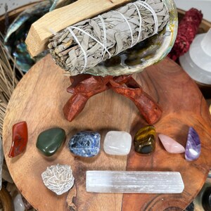 May include: A white sage smudge stick and a wooden palo santo stick are bundled together and placed in a large abalone shell on a wooden stand.  The shell is resting on a wooden surface with a variety of crystals, including red jasper, green aventurine, blue sodalite, clear quartz, tiger's eye, rose quartz, selenite, and amethyst.