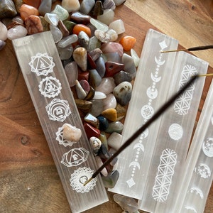 May include: Four white selenite incense holders with different etched designs. The holders are arranged in a row on a wooden surface with a variety of colorful stones scattered around them. A brown incense stick is burning on the holder with the chakra symbols.