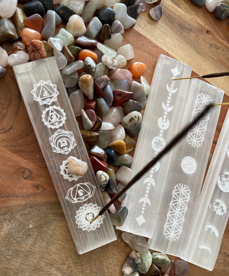 May include: Four white selenite incense holders with different etched designs. The holders are arranged in a row on a wooden surface with a variety of colorful stones scattered around them. A brown incense stick is burning on the holder with the chakra symbols.