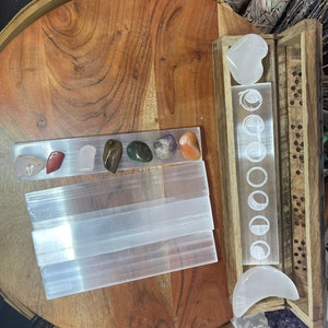 May include: A wooden box with a clear glass insert featuring a moon phase design. The box contains a white selenite heart and a crescent moon. There are also several selenite sticks and various colored crystals on a wooden surface.