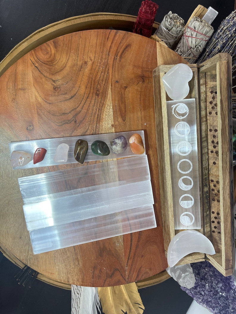 May include: A wooden box with a clear glass insert featuring a moon phase design. The box contains a white selenite heart and a crescent moon. There are also several selenite sticks and various colored crystals on a wooden surface.