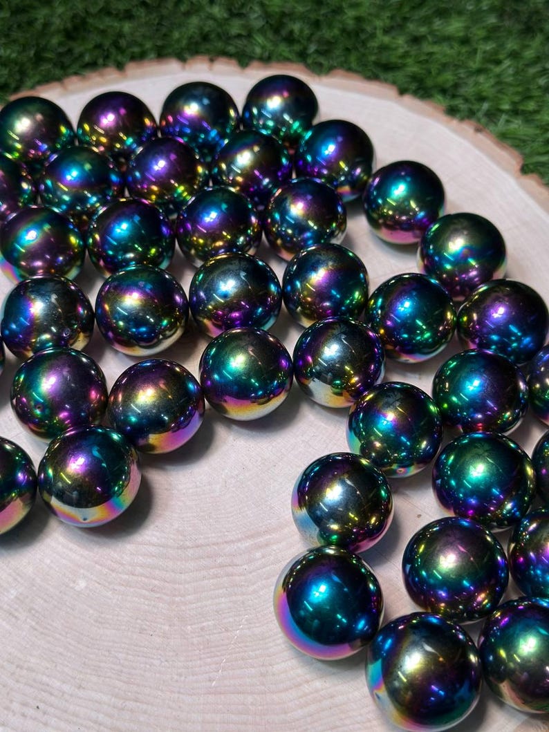 K&ouml;nnte beinhalten: Eine Ansammlung von irisierenden Kugeln mit polierter, reflektierender Oberfl&auml;che. Die Kugeln zeigen eine lebendige Farbpalette, darunter Blau-, Gr&uuml;n-, Lila- und Gelbt&ouml;ne. Sie sind auf einer hellen Holzoberfl&auml;che angeordnet.