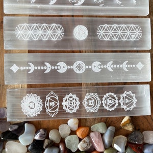 May include: Four white selenite charging wands with various etched designs, including moon phases, geometric patterns, and chakra symbols. The wands are arranged vertically on a wooden surface, with a pile of colorful tumbled stones in the foreground.