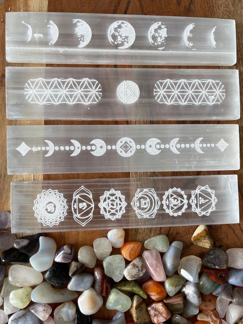 May include: Four white selenite charging wands with various etched designs, including moon phases, geometric patterns, and chakra symbols. The wands are arranged vertically on a wooden surface, with a pile of colorful tumbled stones in the foreground.
