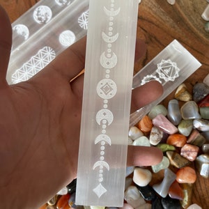 May include: A white selenite wand with a moon and diamond pattern etched into the surface. The wand is surrounded by a variety of colorful stones.