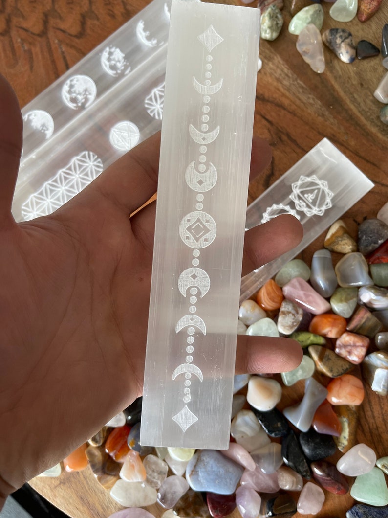 May include: A white selenite wand with a moon and diamond pattern etched into the surface. The wand is surrounded by a variety of colorful stones.